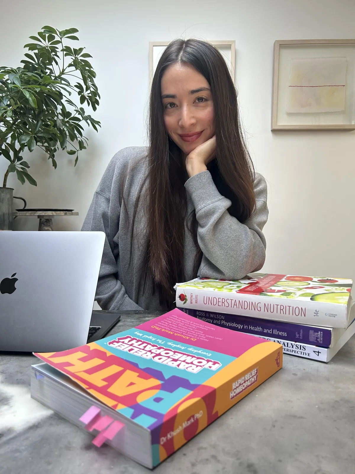 Celine sitting behind table with Apple laptop and nutrition books. In front of the table is Rapid Relief Homeopathy book