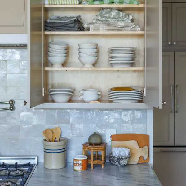 Organized kitchen cabinet with stacked white bowls, plates, and folded linens on wood shelves above a tiled stovetop