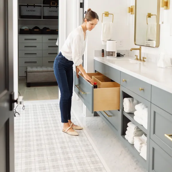 Home organizer opening a bathroom vanity drawer beside open shelves of neatly folded white towels