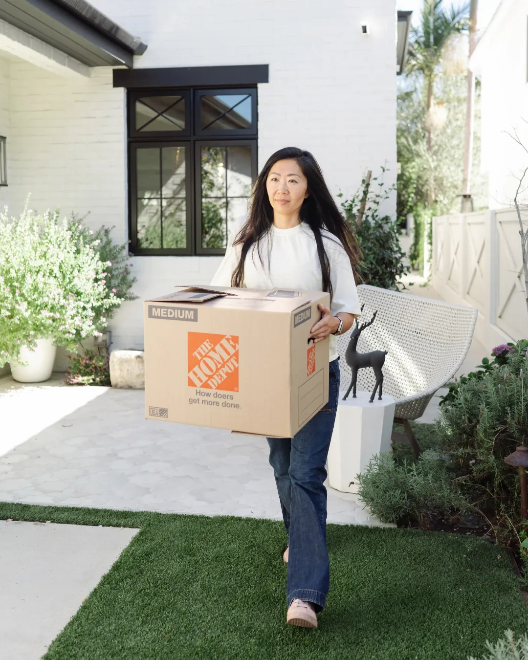 Kaymenity Home organizer carrying a moving box across a front yard toward a white brick home