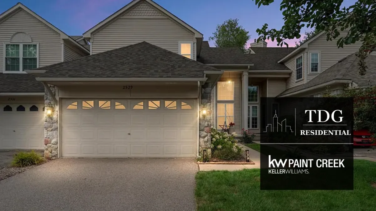 Wixom MI townhouse for sale with a two-story foyer, fireplace, bay window, deck overlooking a wooded backdrop, flexible lower level, and attached two-car garage.