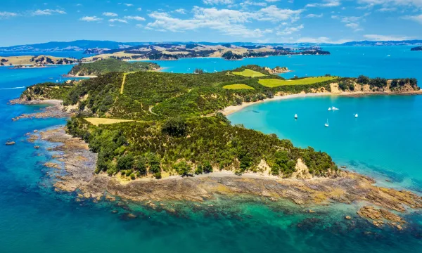 Aerial view of a lush, green island surrounded by crystal-clear turquoise waters and anchored yachts, with rolling hills and distant islands in the background – New Zealand luxury cruise scenery.