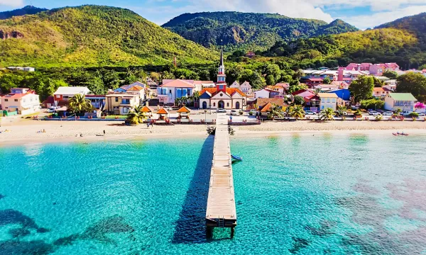 Aerial view of a colorful Caribbean seaside village with a church, sandy beach, wooden pier, turquoise water, and lush green hills in the background—Martinique.