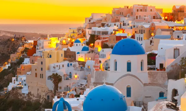 Sunset over the iconic blue-domed churches and whitewashed buildings of Oia, Santorini, glowing with warm golden light on the cliffs of the Aegean Sea.