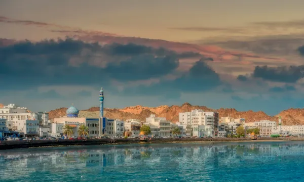 Stunning view of Muscat’s waterfront at sunset, with traditional white buildings, a mosque, and dramatic mountains rising in the background – a serene Arabian Gulf gem featured on Middle East cruises with Luxury Sassy Cruises.