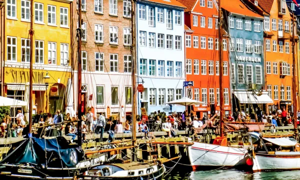 A lively scene from Nyhavn, Copenhagen, with iconic colorful buildings, moored boats, and people enjoying outdoor cafés by the canal.