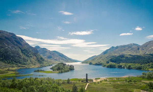 A panoramic view of the Scottish Highlands with a serene lake surrounded by lush green mountains and a historic stone monument at the water’s edge under a clear blue sky.