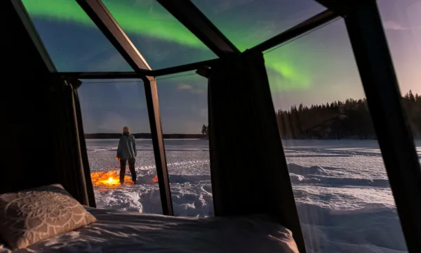 View from a glass igloo in a snowy wilderness, with a person standing outside under the glowing green aurora borealis lighting up the night sky.