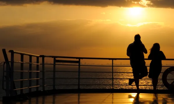 Two people stand on the deck of a cruise ship at sunset, gazing out over the ocean with the distant UK coastline visible on the horizon, capturing the anticipation and beauty of cruising from the UK.