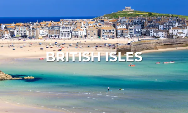 A picturesque seaside town with whitewashed houses overlooks a sandy beach and turquoise waters in the British Isles. Small boats are anchored in the bay, with green hills and a blue sky in the background. White text reads "British Isles."