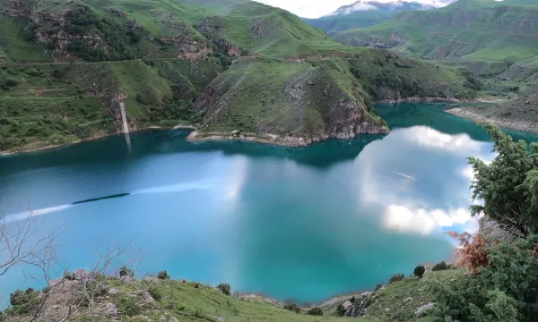 A panoramic view of a turquoise-blue fjord surrounded by steep green hills in Patagonia, reflecting a partly cloudy sky in its calm waters.