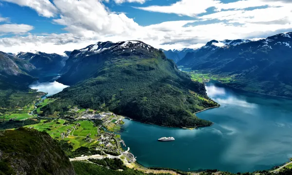 A panoramic aerial view of a deep, turquoise-blue fjord flanked by steep, green mountains and a small village on the shoreline, with a white cruise ship gliding across the calm water under a partly cloudy sky.
