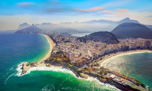 Aerial view of Rio de Janeiro’s coastline at sunrise, showing the curved sands of Copacabana Beach, high-rise cityscape, and misty Sugarloaf Mountain in the distance