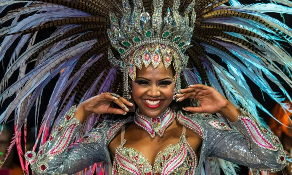 A smiling samba dancer in a glittering silver and pink costume with an elaborate feathered headdress, posing under colorful lights at a Brazilian Carnival parade
