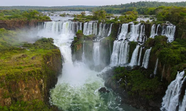 A wide-angle view of the multi-tiered Iguazú Falls plunging into a misty gorge, surrounded by lush green rainforest.