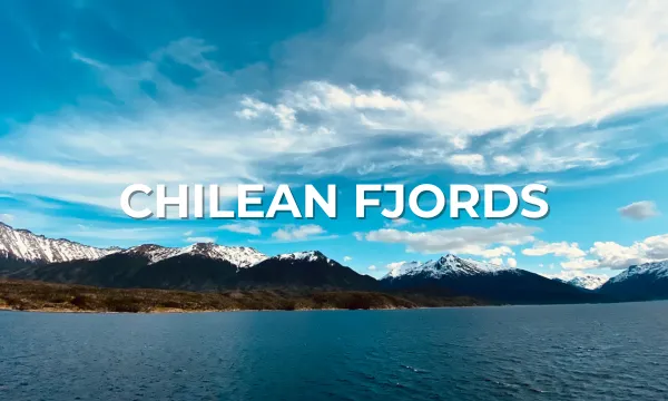 Wide expanse of calm, deep-blue fjord water in southern Chile, framed by rugged snow-capped peaks under a bright, partly cloudy sky.