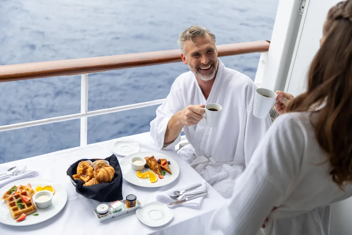 Couple enjoying gourmet breakfast in robes on a private cruise balcony overlooking the ocean.