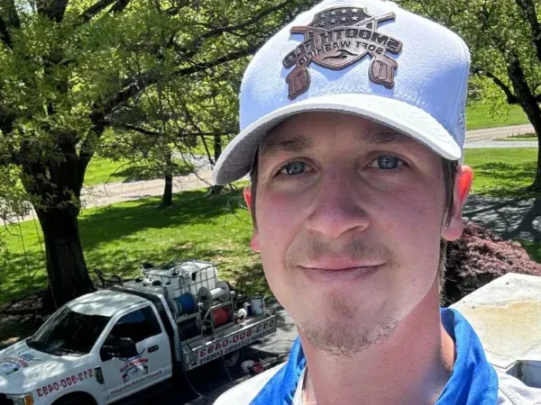 A man wearing a white baseball cap with a company logo takes a selfie with a white soft washing truck in the background.