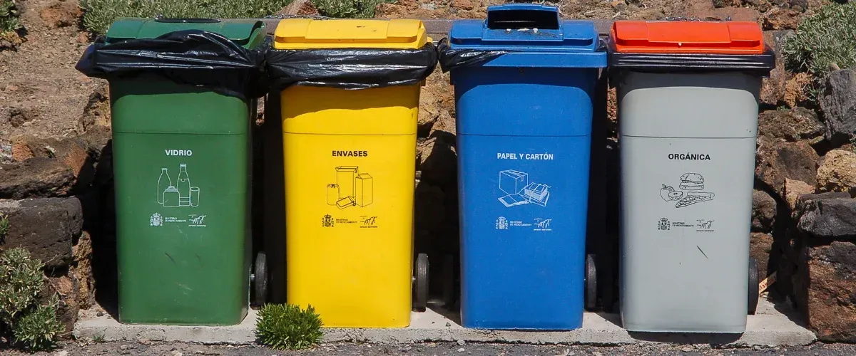 Four colorful recycling bins, green, yellow, blue, and gray, standing in a line outdoors on a sunny day.