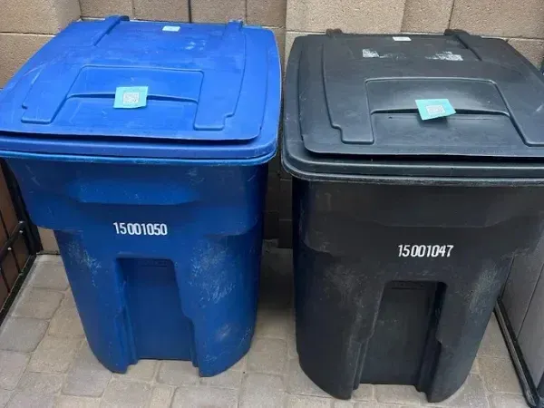 Two outdoor garbage cans, one blue and one black, with QR code stickers on their lids and numbers on their sides.