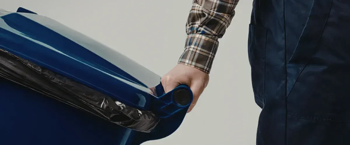 A person in a plaid shirt and dark overalls holds the corner of a dark blue recycling bin with a black trash bag inside.
