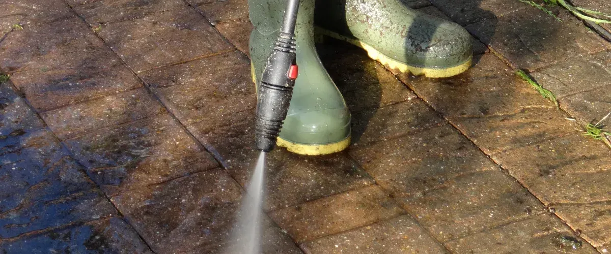 A person in green wellington boots using a power washer to clean dirty, brown patio pavers.