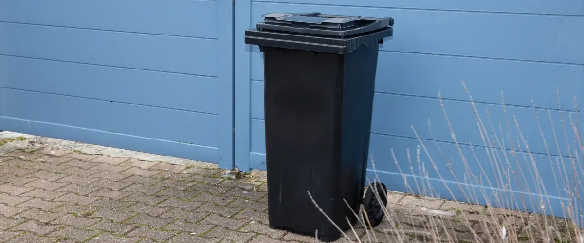 A black plastic waste bin with wheels stands upright on a brick patio next to a blue horizontal slat gate.