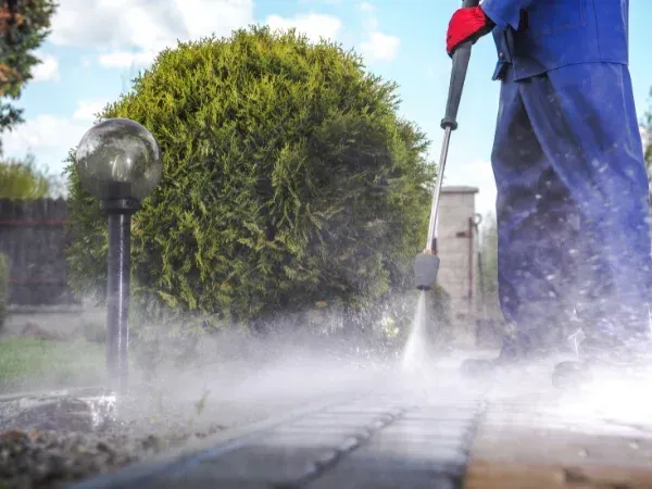 A person in blue overalls and red gloves pressure washing a stone garden walkway in a close-up, low-angle shot.