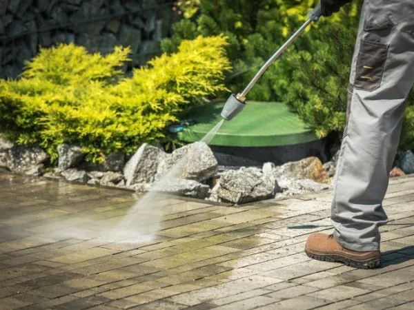 A person in work pants and boots power washing a dirty brick patio with a high-pressure stream of water.