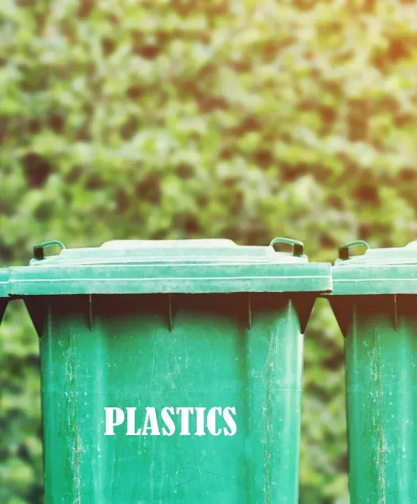 A close-up view of a dark green plastic recycling bin with a metal handle, positioned outdoors on grass next to pavement.