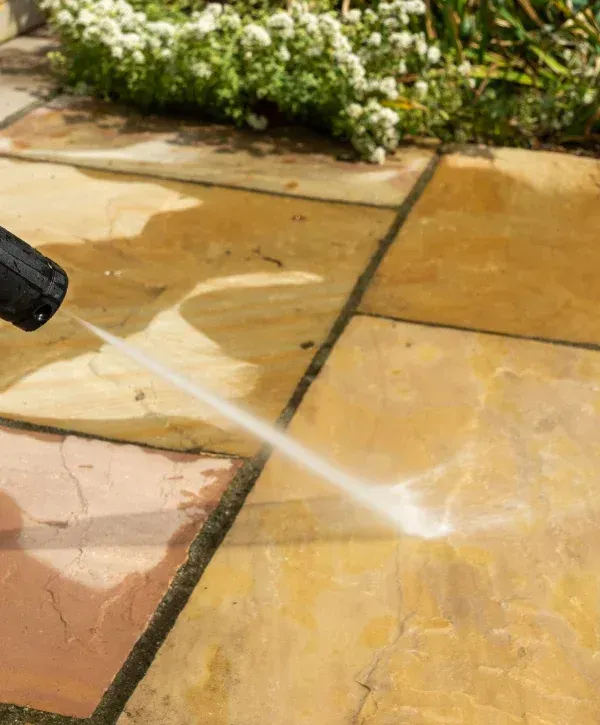 A power washer cleaning dirty patio slabs, with a jet of water visibly removing grime from the light brown paving.
