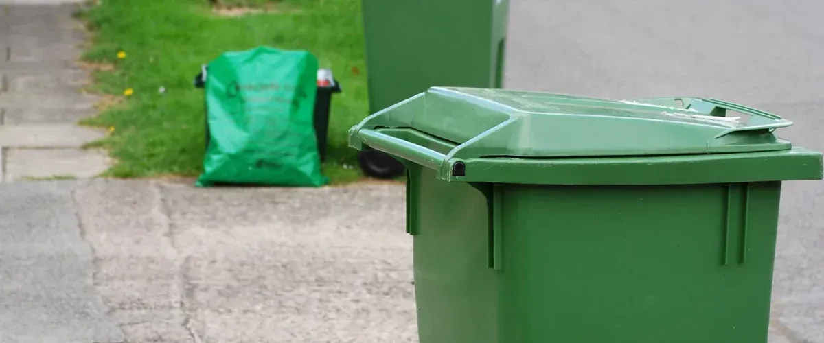 A close-up of a green recycling bin lid with a green bag and another bin in the blurry background on a sidewalk.