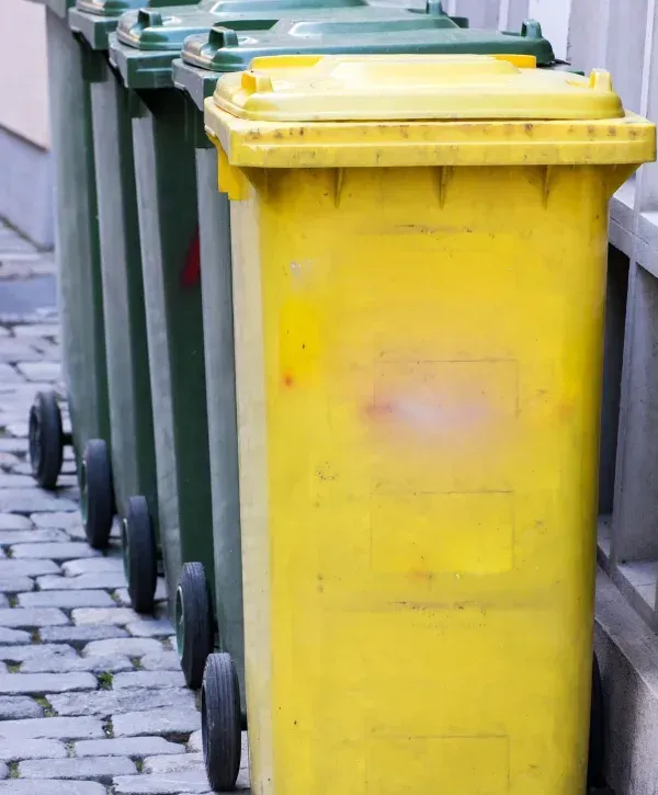 A yellow and several green wheeled garbage bins lined up on a cobblestone path next to a building.