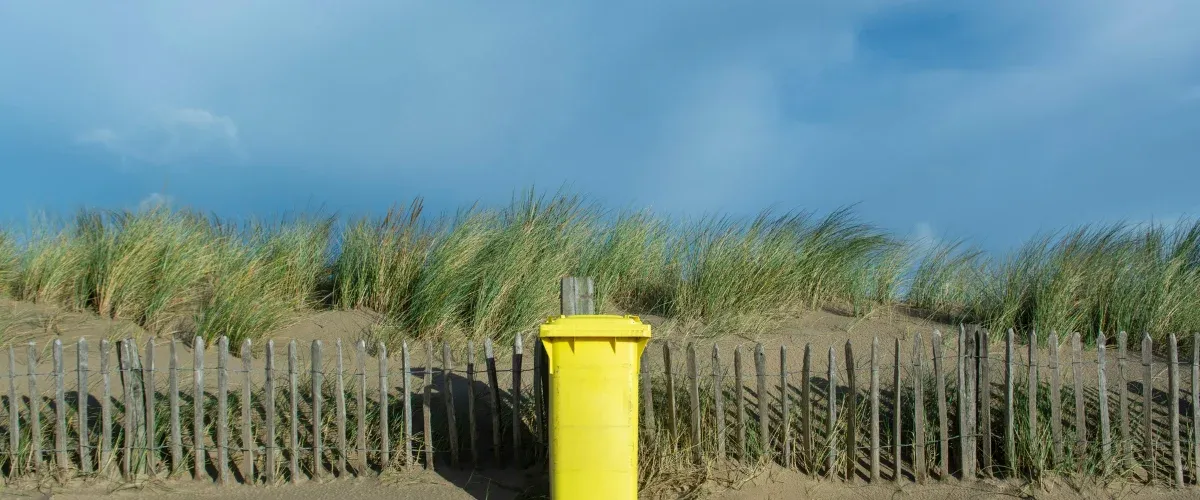 A bright yellow recycling bin stands in front of a wooden fence, sand dunes with green grass, and a blue sky.