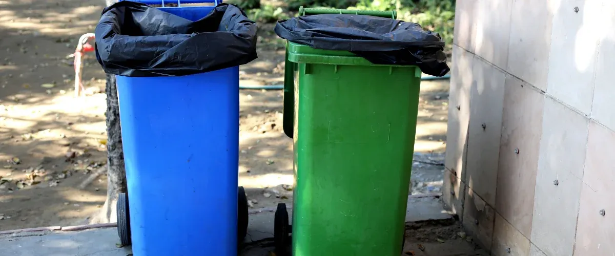 A row of blue and black trash and recycling bins lined up next to a paved street on a sunny day.