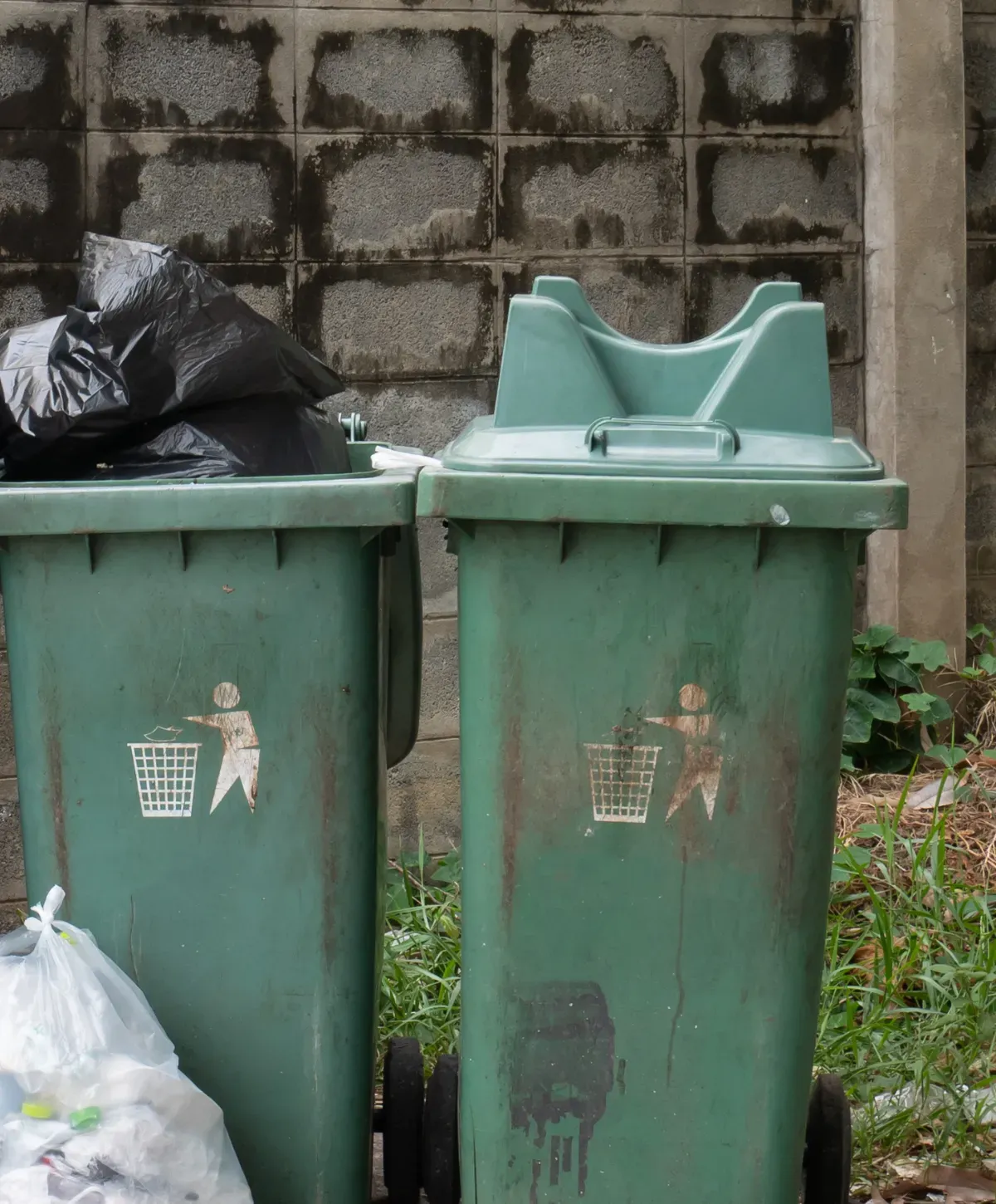 Two dirty green garbage bins with trash symbols stand against a concrete block wall, one overflowing with black trash bags.