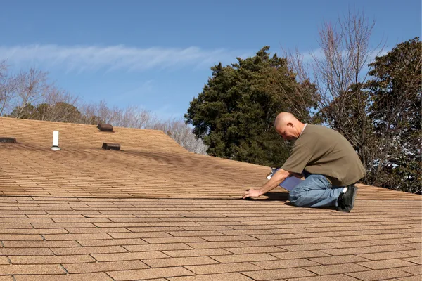 Home inspector kneeling on a roof examining shingles under a clear sky during an Orlando wind mitigation inspection.