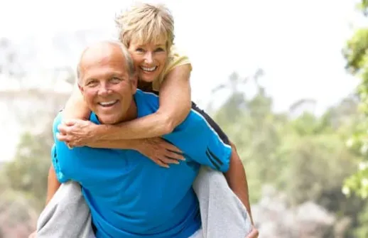 Smiling senior couple outdoors, woman on man’s back, highlighting discussion on whether vitamin D is best received as injections or oral supplements.