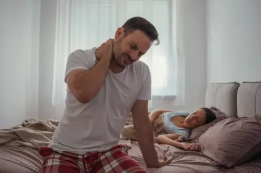 Man sitting on the edge of a bed holding his neck in pain while a woman sleeps in the background, symbolizing chronic pain and the need for non-opioid, non-pharmaceutical treatment options.