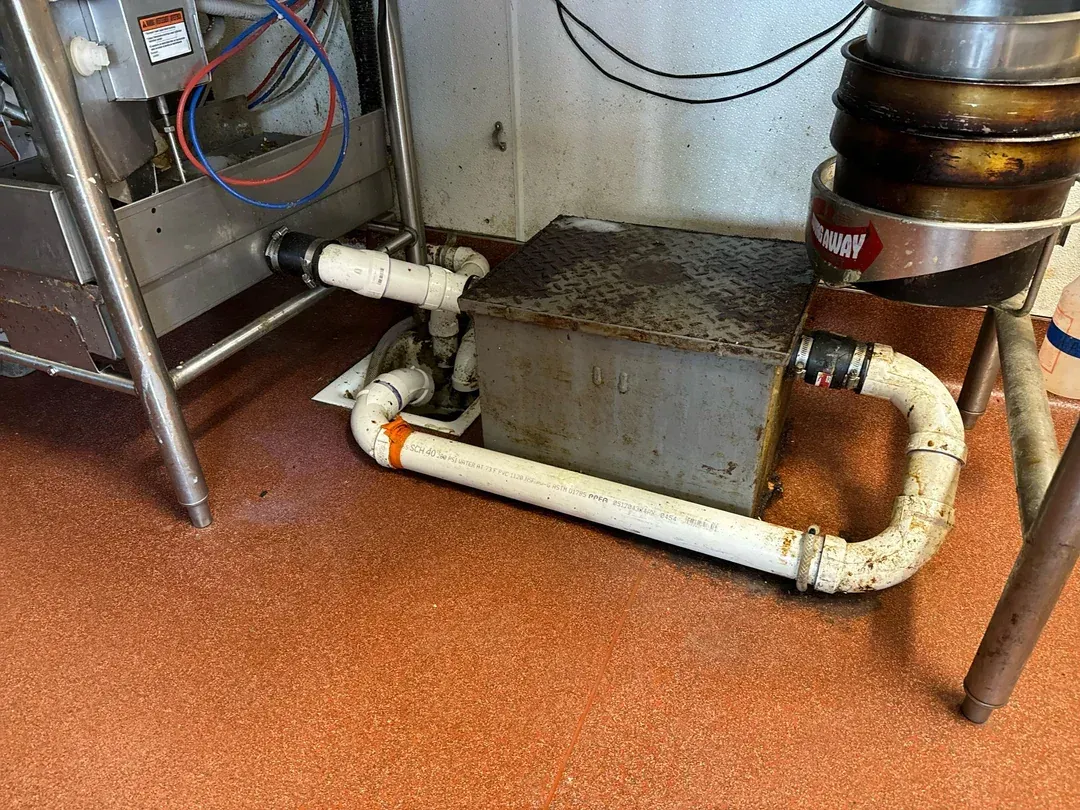 Overflowing commercial grease trap with visible buildup and floor stains near kitchen equipment.
