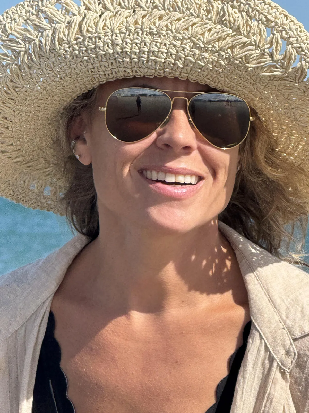 Kandice smiling in a straw hat and sunglasses on the beach.