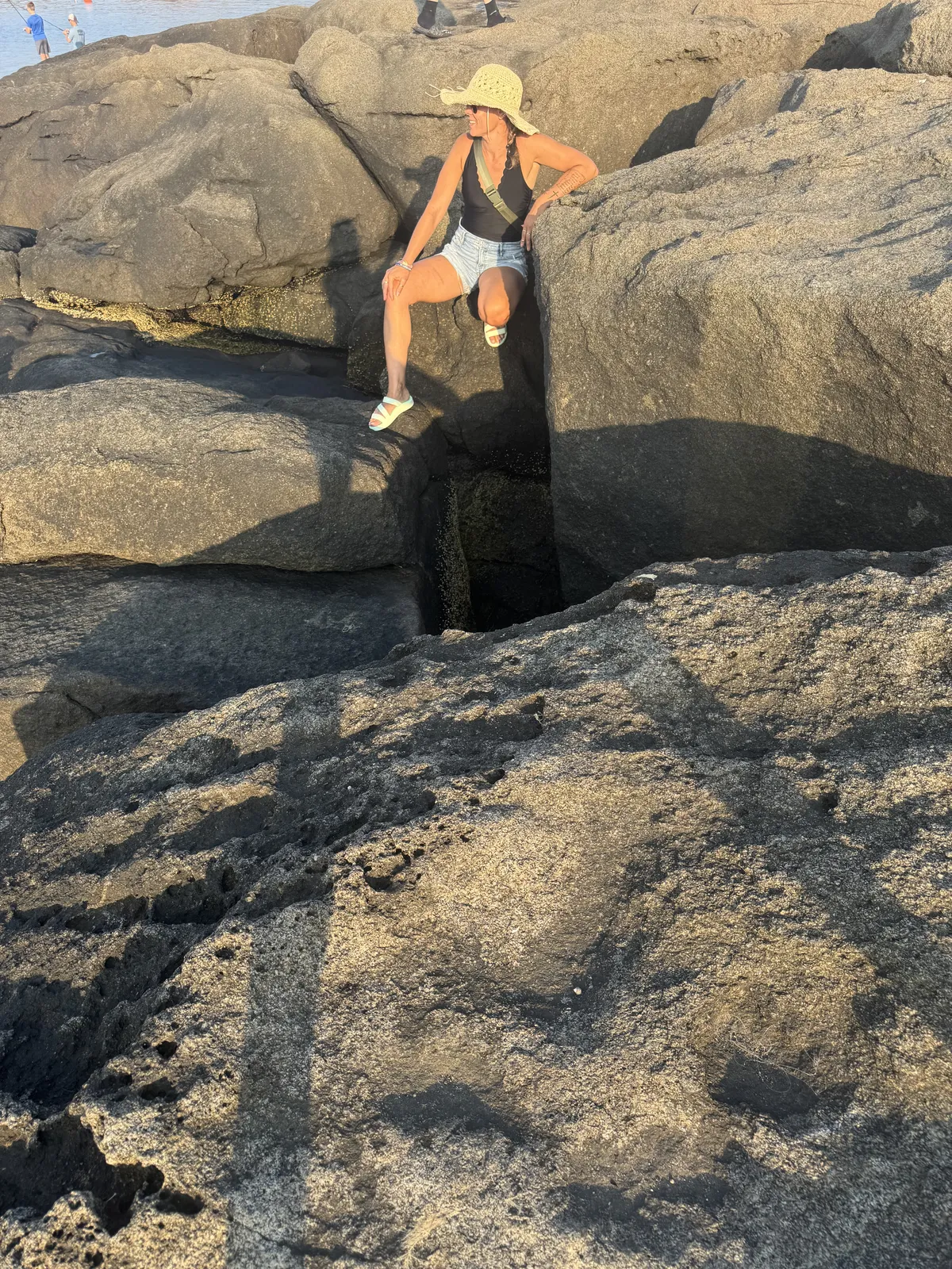 Kandice sitting on large rocks by the ocean in a sun hat.