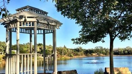 A white wooden gazebo overlooking the calm waters of Atascadero Lake, surrounded by green trees on a sunny day.