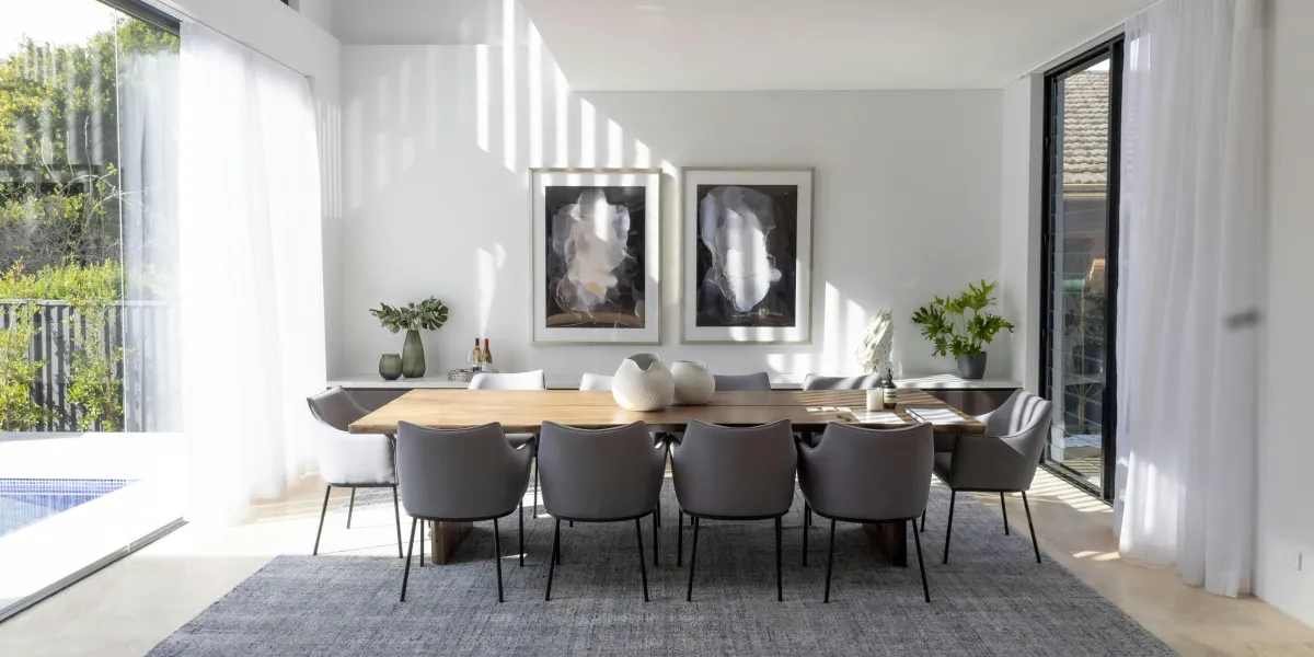 Sunlit dining room with a sculptural wood table, upholstered chairs, and floor-to-ceiling glass overlooking a pool