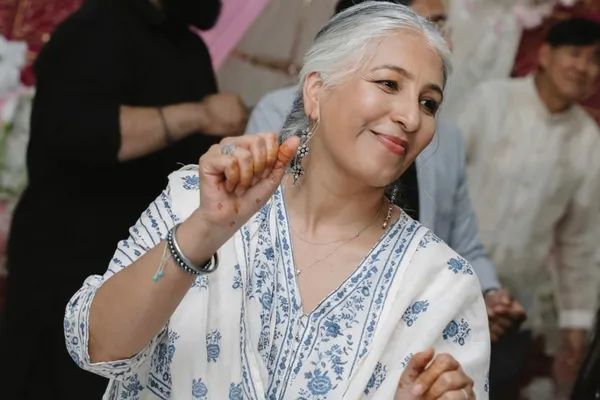 A woman smiling and dancing at a celebration, wearing a white and blue floral outfit with mehndi on her hands.