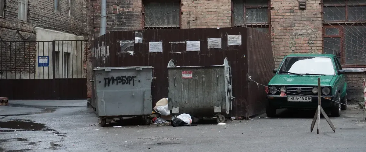 A dirty, neglected alleyway featuring two overflowing dumpsters, a run-down green car, and brick buildings.