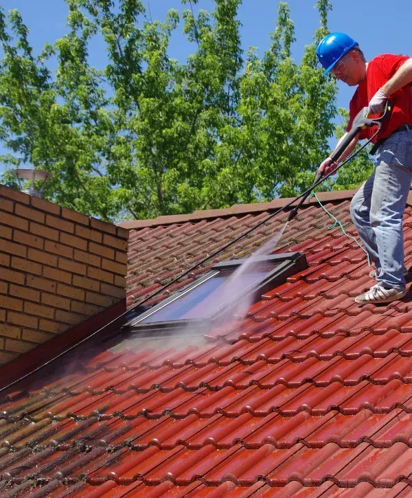 A man in a blue hard hat and red shirt power washes a red tiled roof with a skylight on a sunny day.