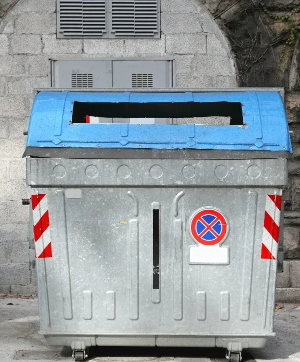 A large grey metal waste container with a blue plastic lid and a no parking sign on its front.
