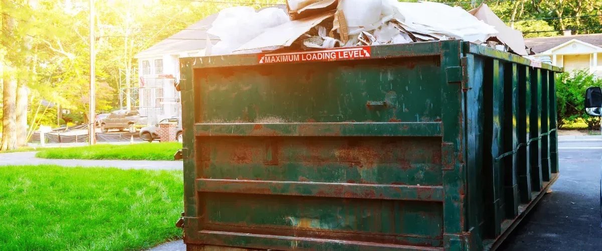 A large, dark green dumpster overflowing with construction debris and cardboard on a sunny day.