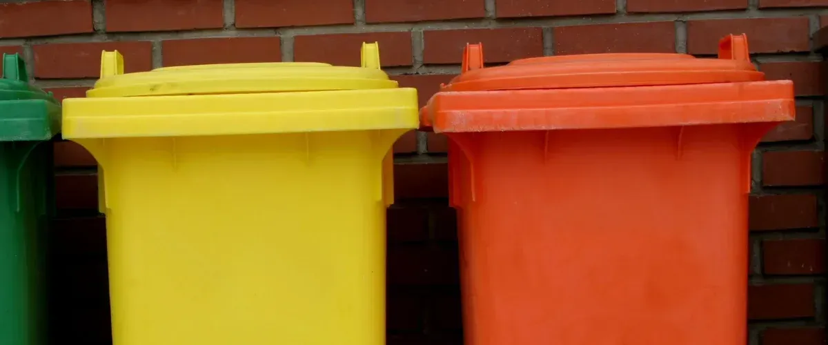 A close-up of green, yellow, and orange plastic garbage bins lined up against a red brick wall.